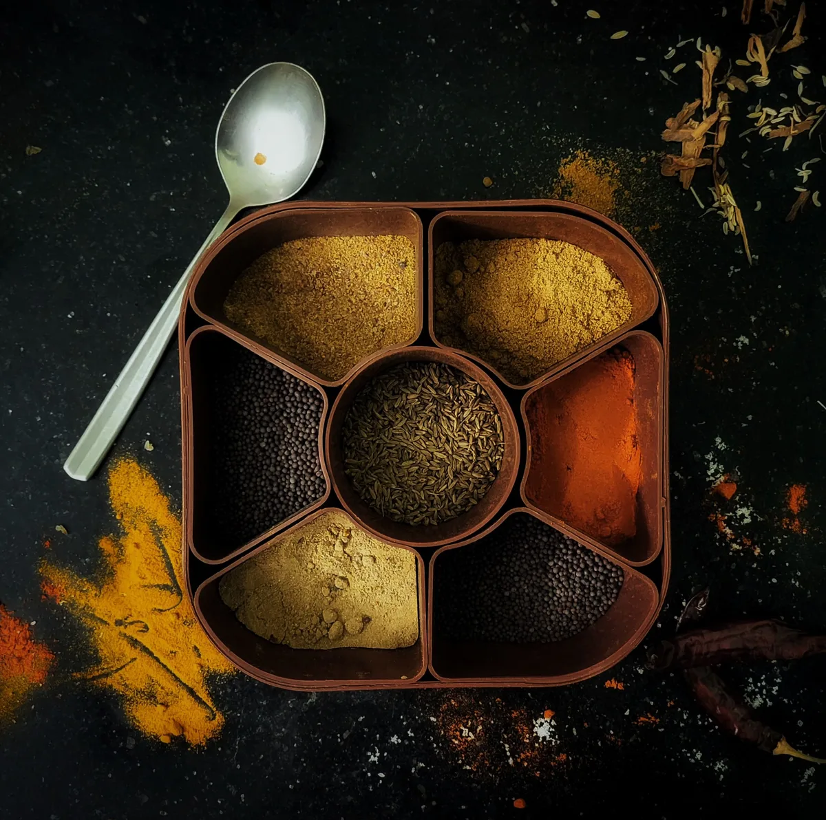 A spice box with whole and ground spices in its compartments, a spoon beside it