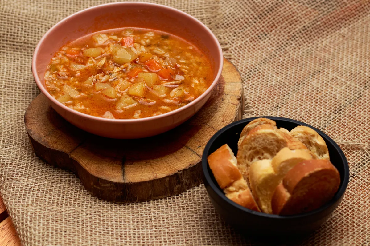 A bowl of vegetable soup on a wooden board with a bowl of bread on the side