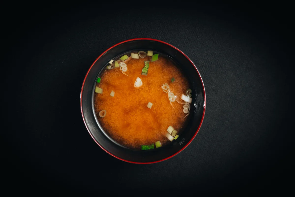 A black bowl of miso soup topped with sliced green onion, seen from above on a dark table