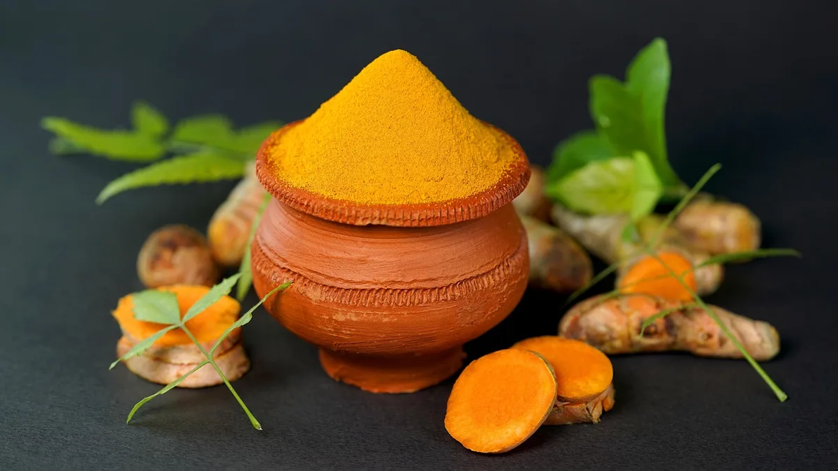 A clay pot heaped with ground turmeric, surrounded by fresh turmeric roots, cut slices and green leaves