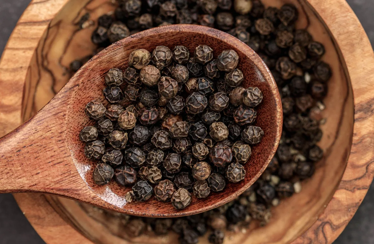A wooden spoon full of black peppercorns resting in a wooden bowl of more peppercorns