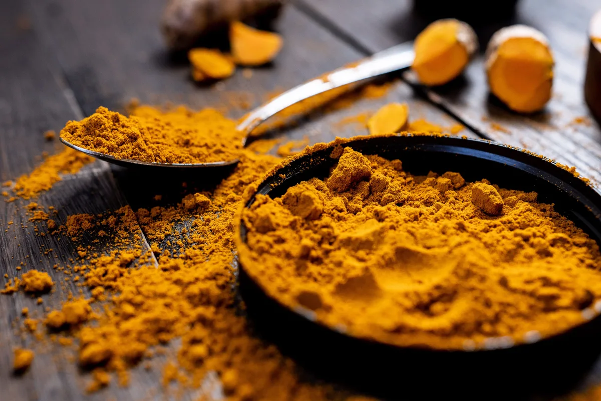 Ground turmeric heaped in a dark dish and spilling from a spoon, with cut pieces of fresh turmeric root behind