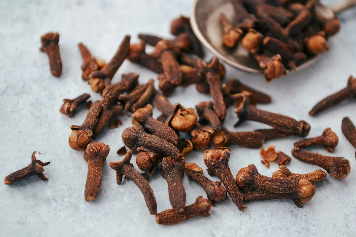 Whole cloves scattered on a gray counter, with more in a metal spoon