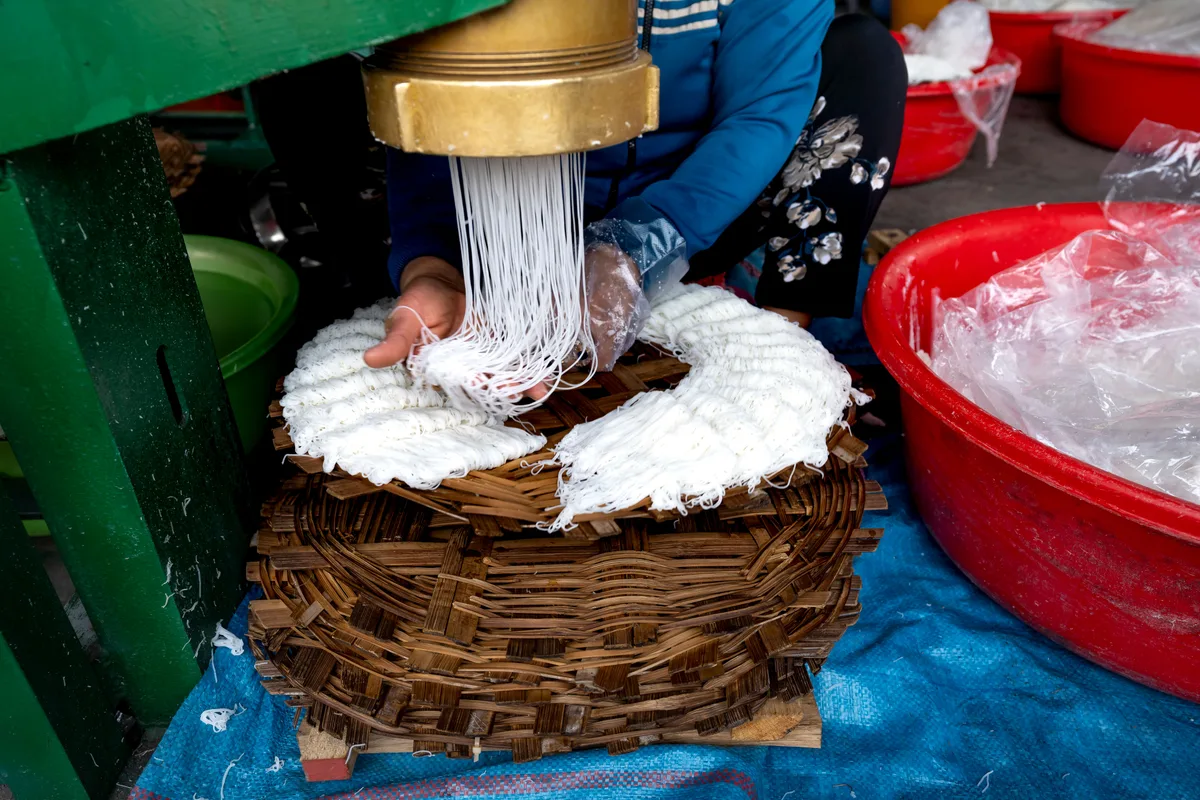 Thin white noodles pressed out of a brass extruder and coiled into a round basket