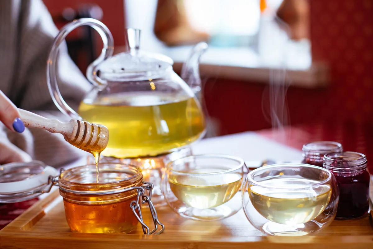 Honey drips from a wooden dipper into a jar beside a glass teapot of hot tea