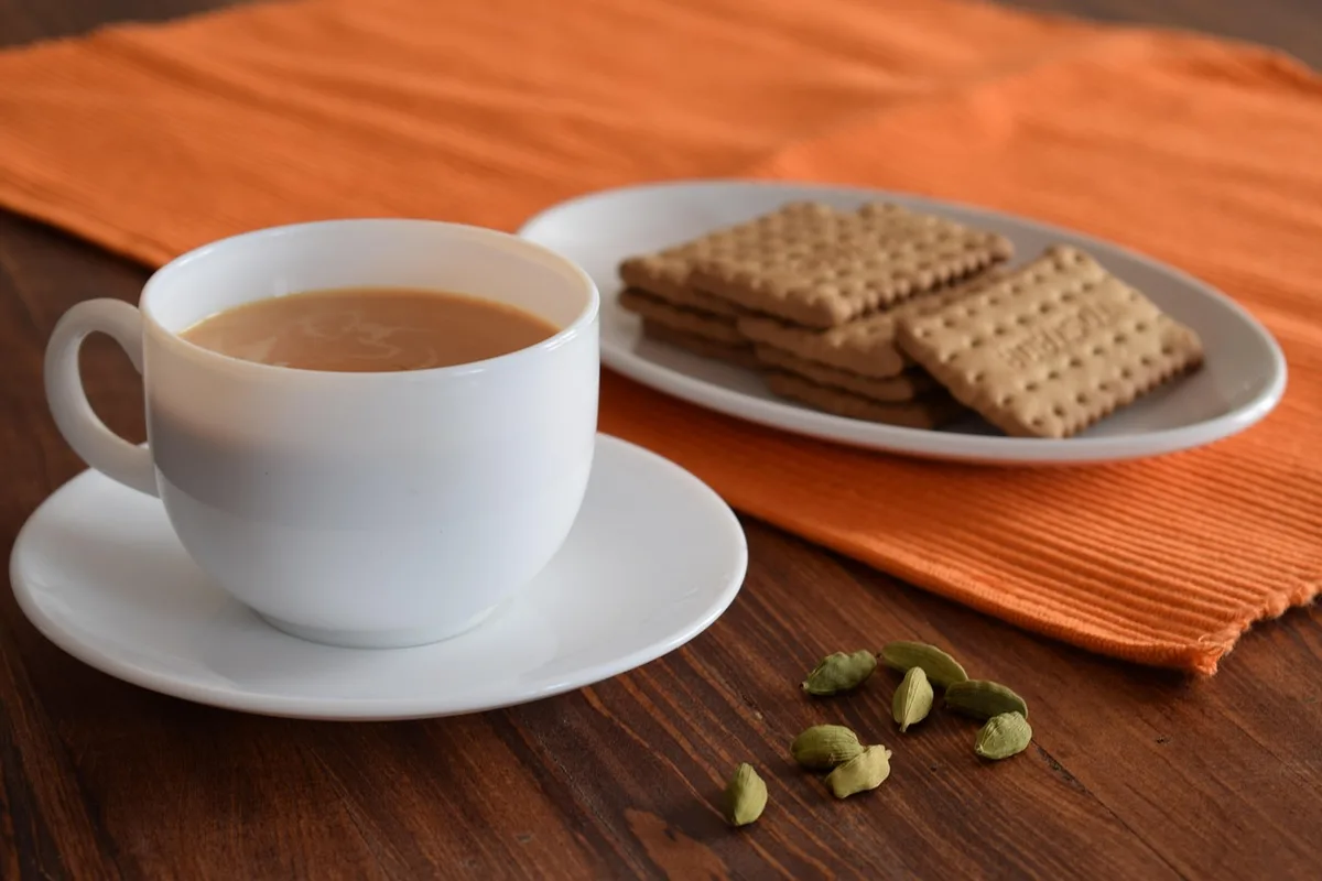 A cup of milky tea with a plate of biscuits and a few green cardamom pods on the table