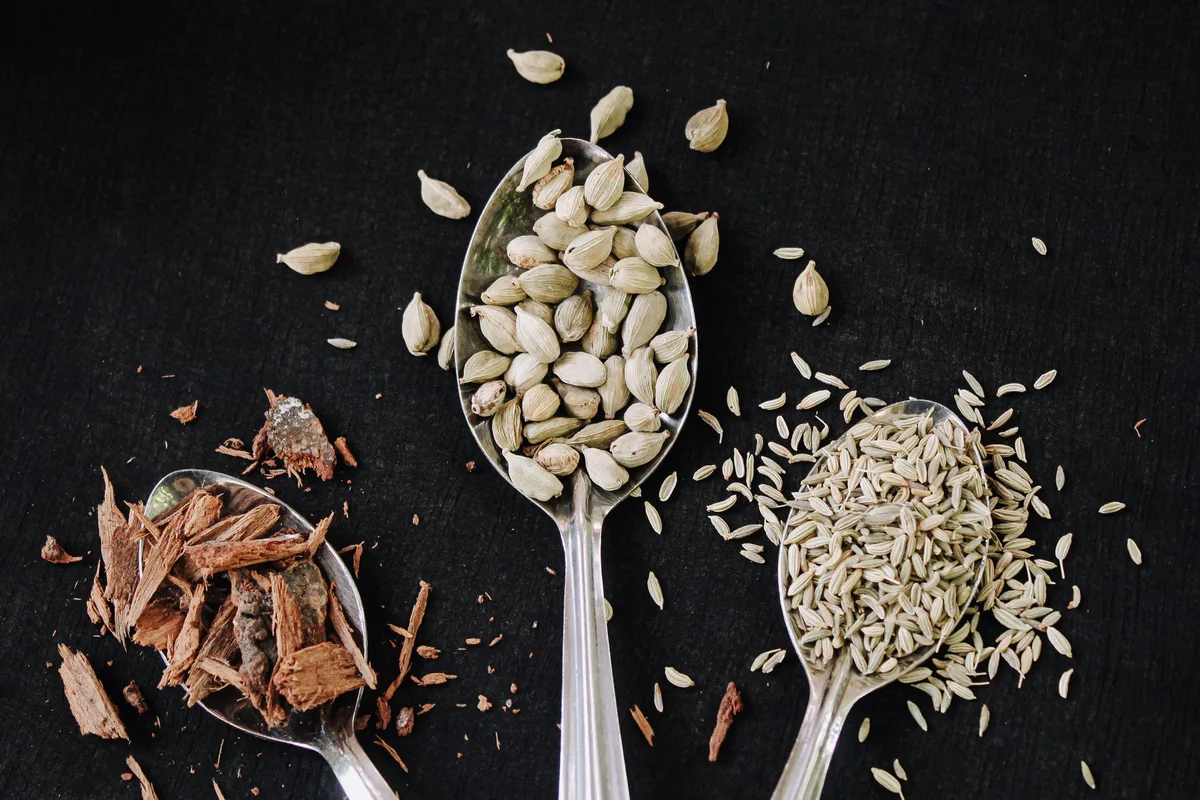 Three spoons of whole spices on a dark cloth: cardamom pods, small pale seeds and broken pieces of bark