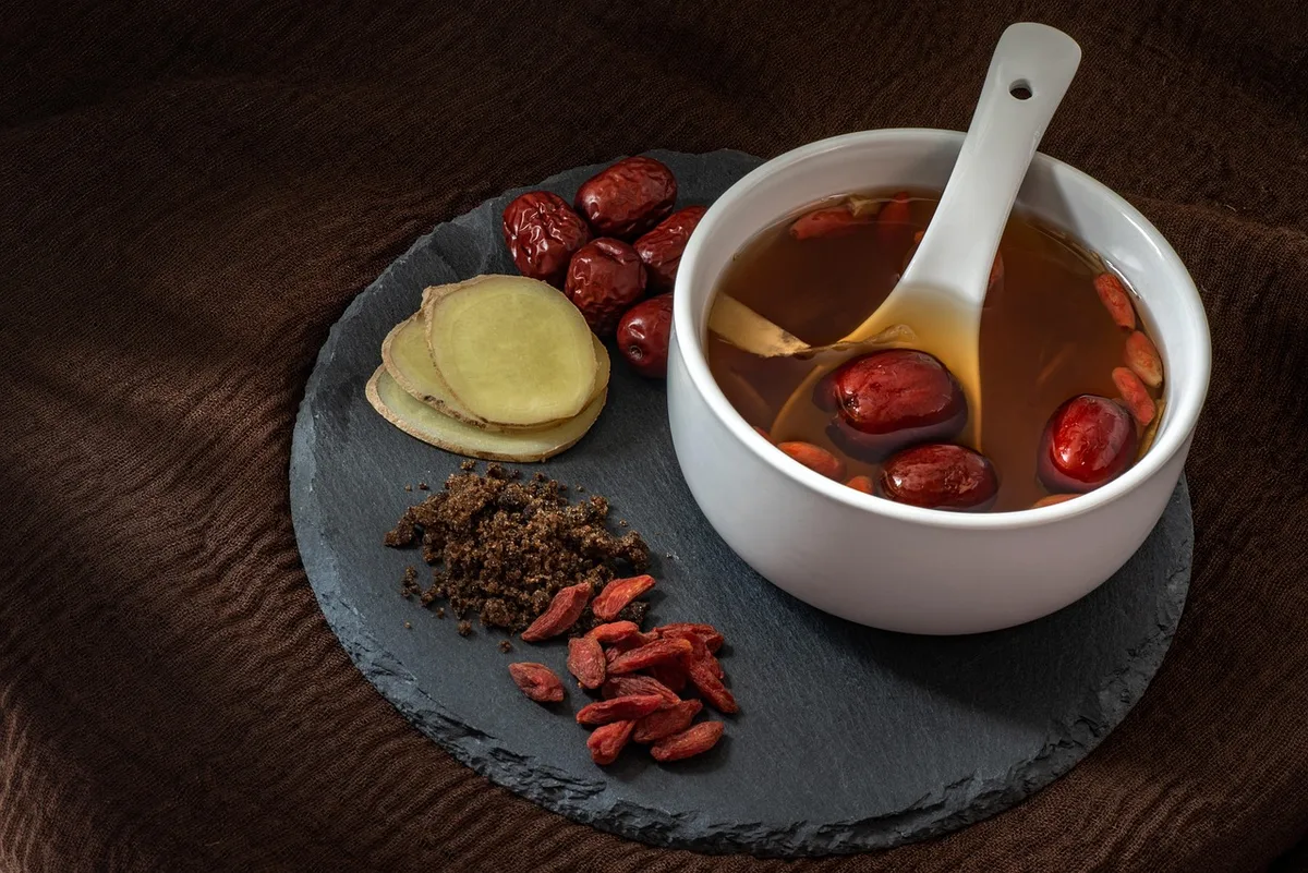 A bowl of amber red date tea with a spoon, next to dried jujubes, sliced ginger and goji berries on a slate board
