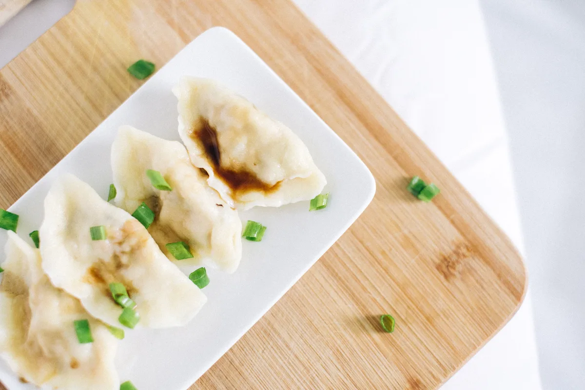 Boiled dumplings on a white plate with a dark dipping sauce and chopped scallions
