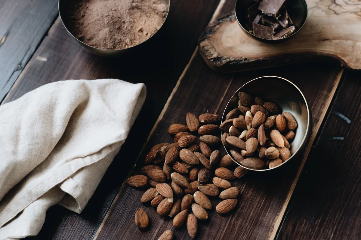 Almonds spilling from a small metal bowl onto a dark wooden board, with cocoa powder and chocolate pieces behind