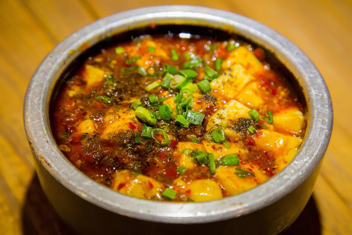 Mapo tofu in a metal pot, the tofu in red chilli oil topped with scallions and ground Sichuan pepper