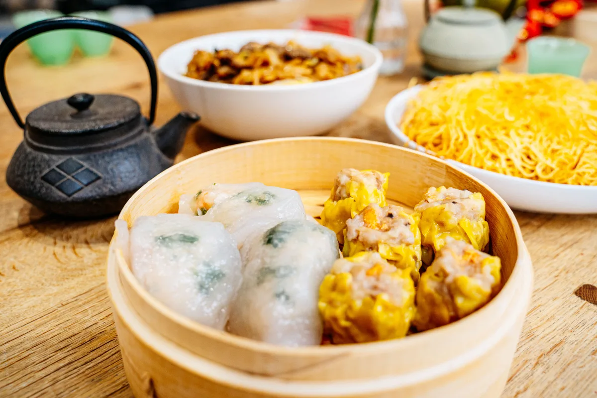 A bamboo steamer of siu mai and translucent steamed dumplings, with a cast iron teapot and noodles behind