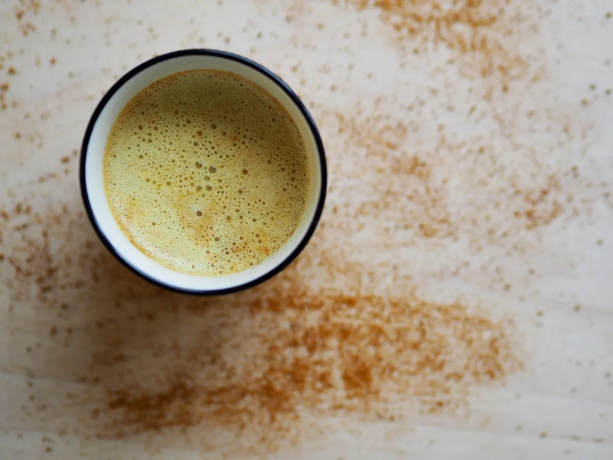 A cup of golden turmeric milk seen from above, with ground spice scattered across the table