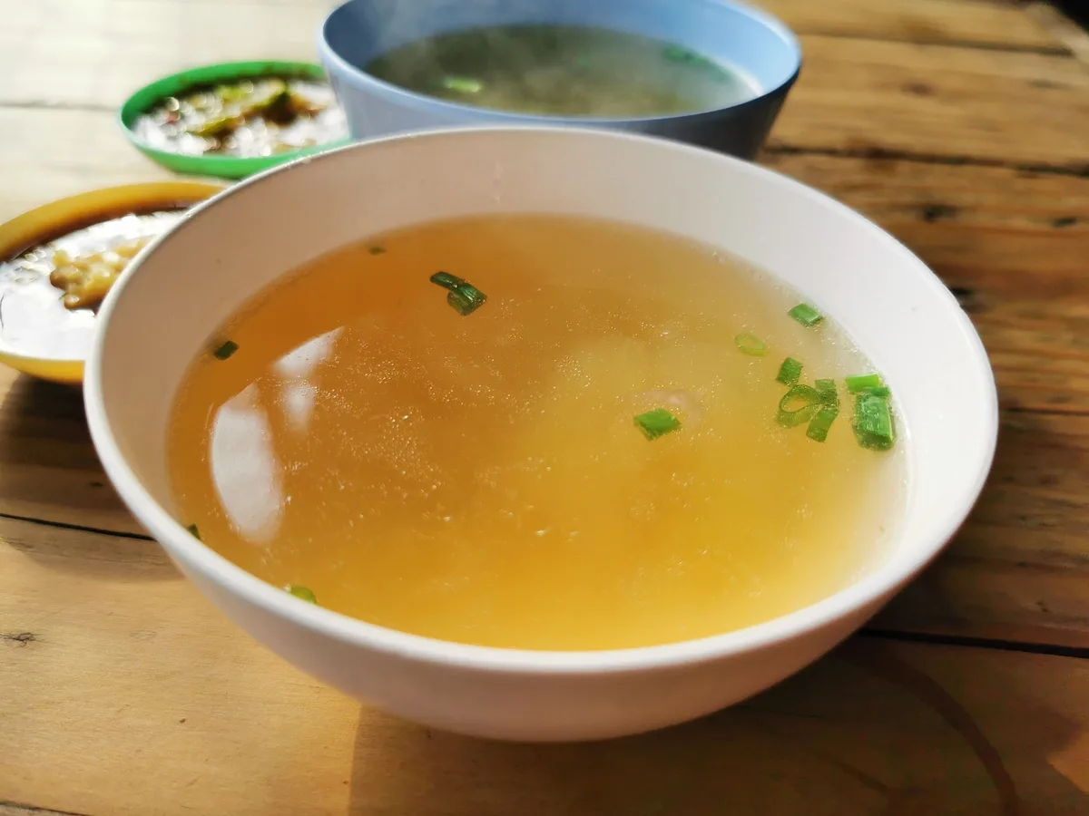 A white bowl of clear golden broth topped with sliced scallions