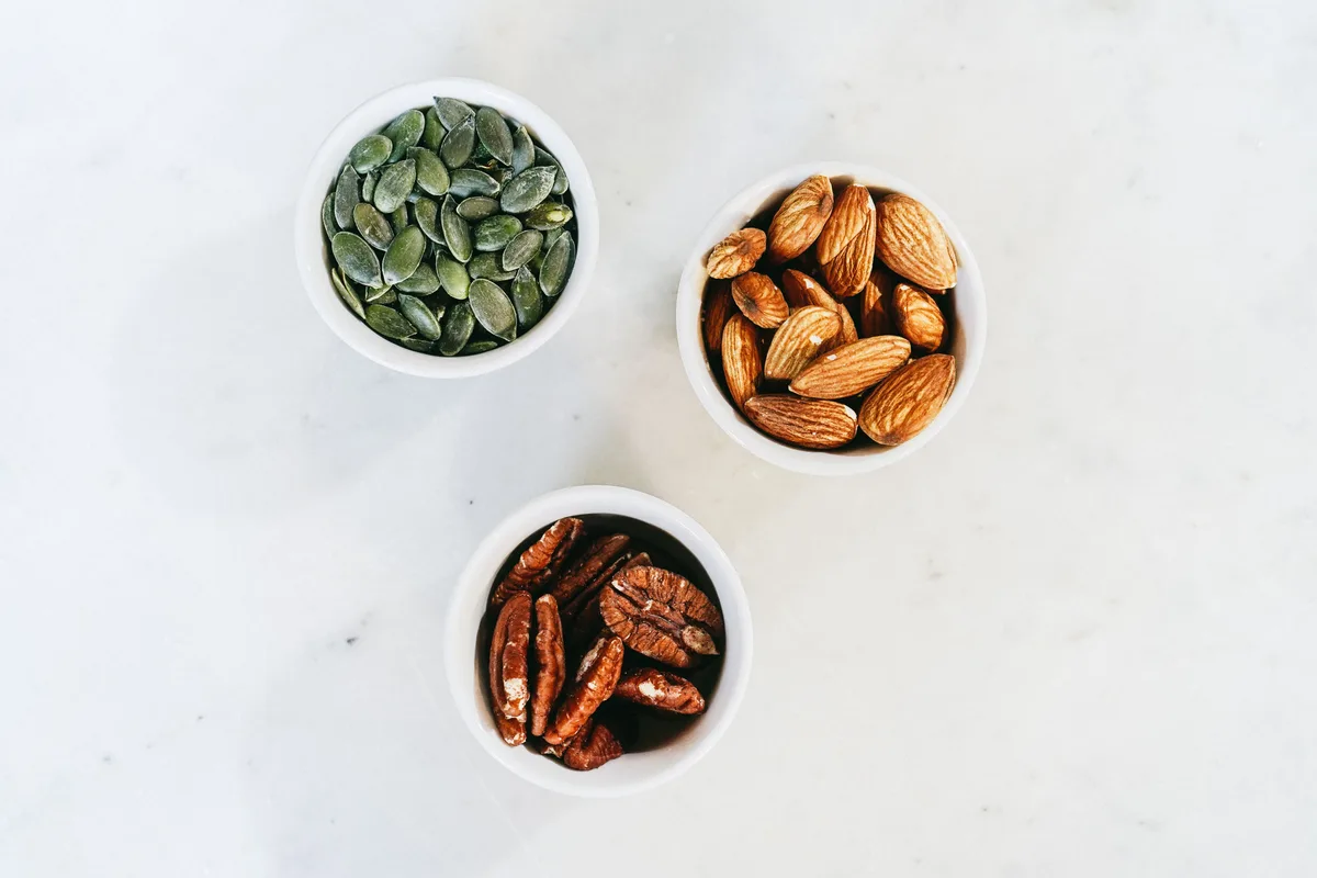 Three small bowls of pumpkin seeds, almonds and pecans on a white table