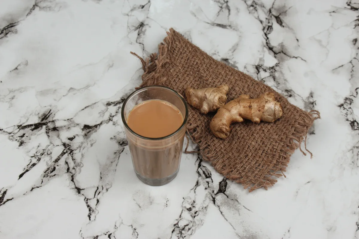 A glass of milky chai on a burlap mat with fresh ginger root, on a marble counter