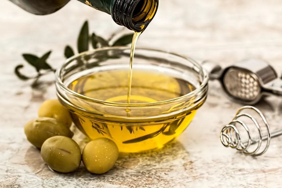 Olive oil poured from a bottle into a glass bowl, green olives beside it
