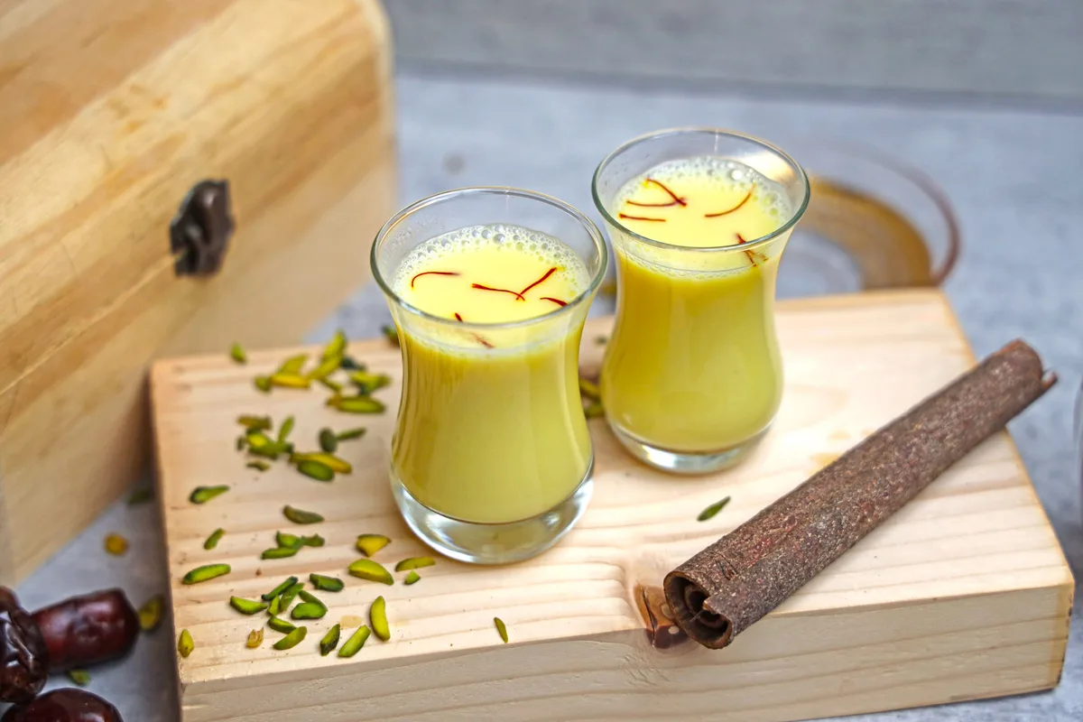 Two small glasses of saffron milk with pistachios and a cinnamon stick on a wooden board