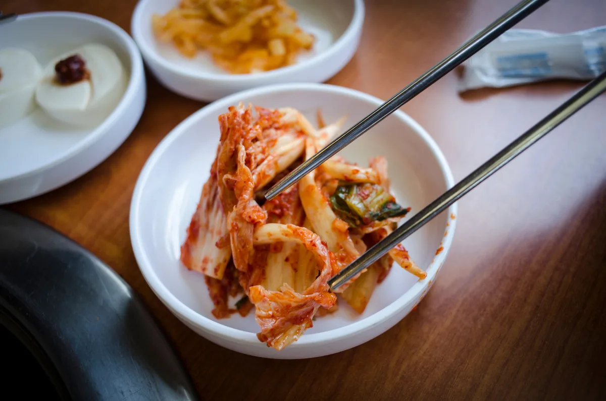 Chopsticks lifting kimchi from a white bowl, with other small side dishes around it