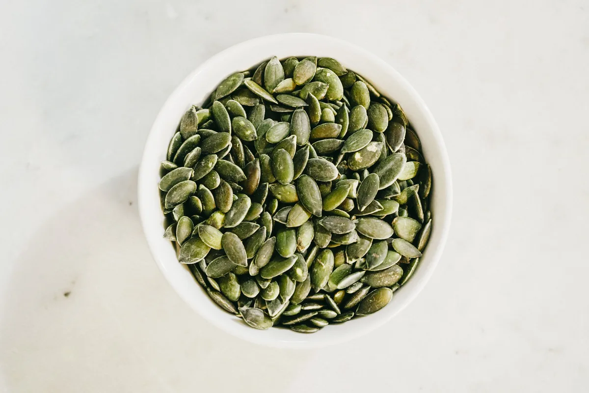 A white bowl of green hulled pumpkin seeds on a marble counter