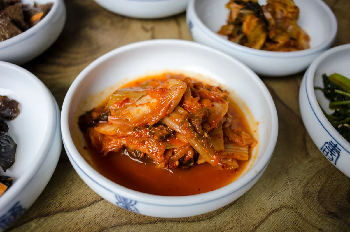A white bowl of napa cabbage kimchi in red chili brine, surrounded by small side dishes