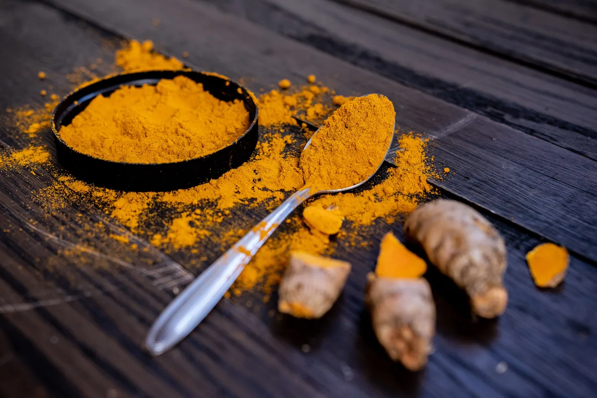 Ground turmeric in a small dish and on a spoon, with fresh turmeric roots on a dark wooden table