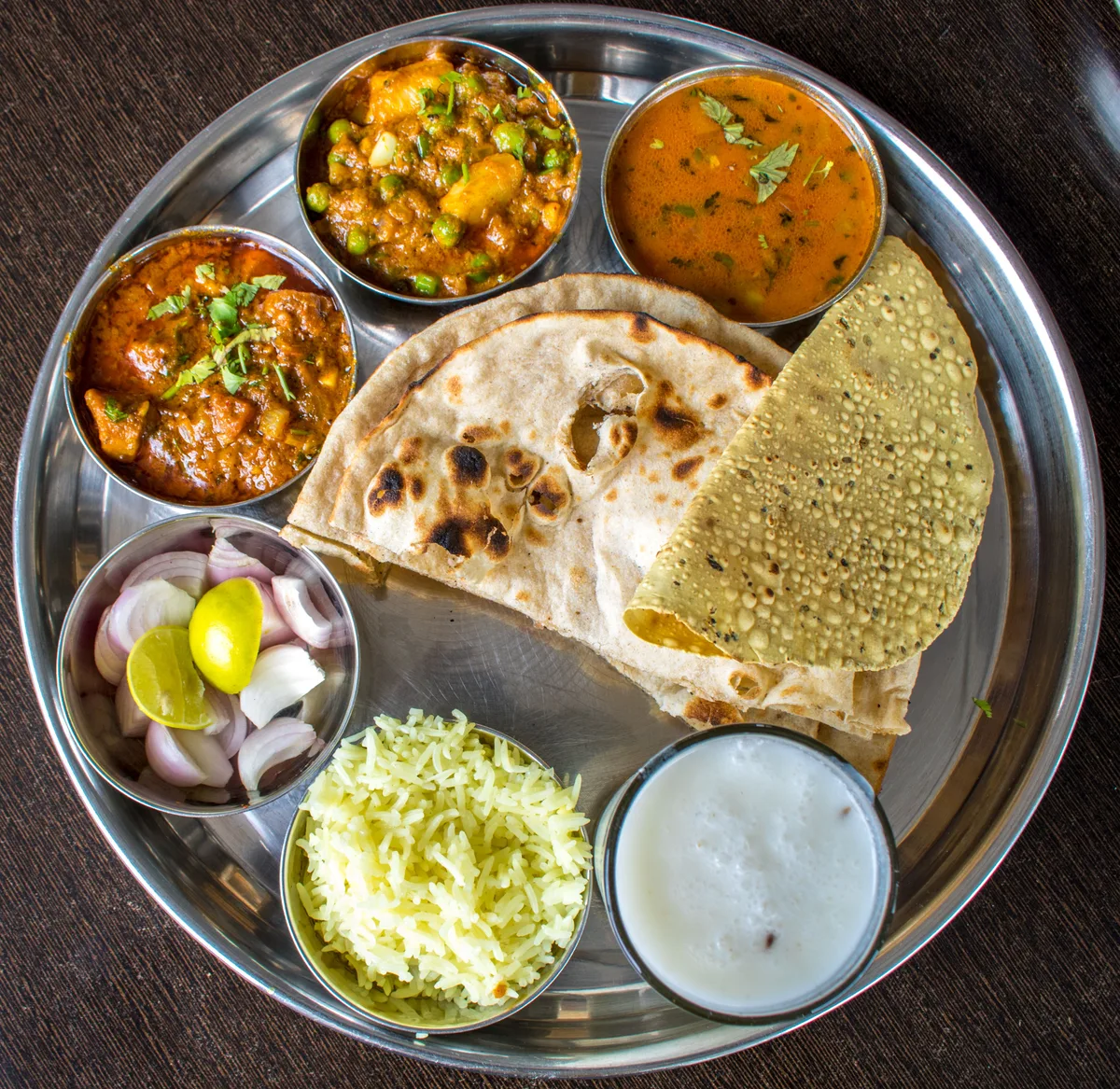 A steel thali with curries, naan, papad, sliced onion, shredded cabbage and yogurt