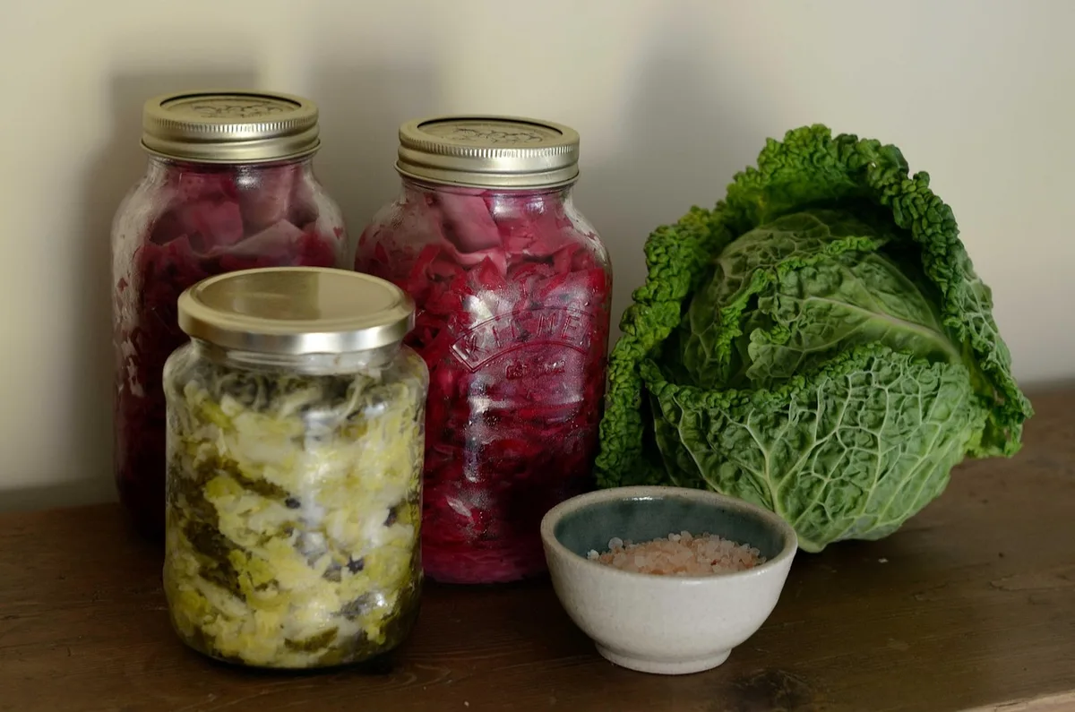 Jars of fermented white and red cabbage beside a whole cabbage and a small bowl of salt