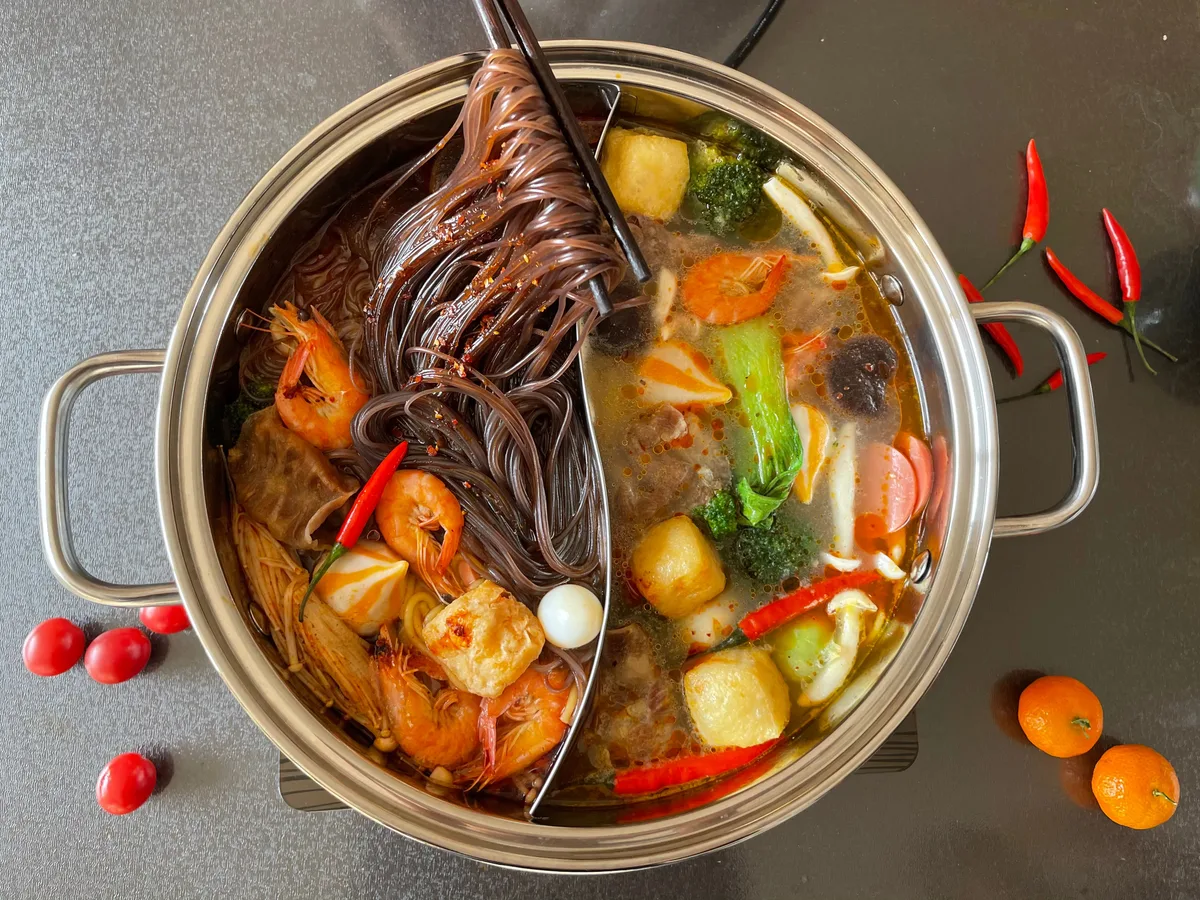 A divided hot pot with two broths, filled with prawns, noodles, potatoes, broccoli and chillies