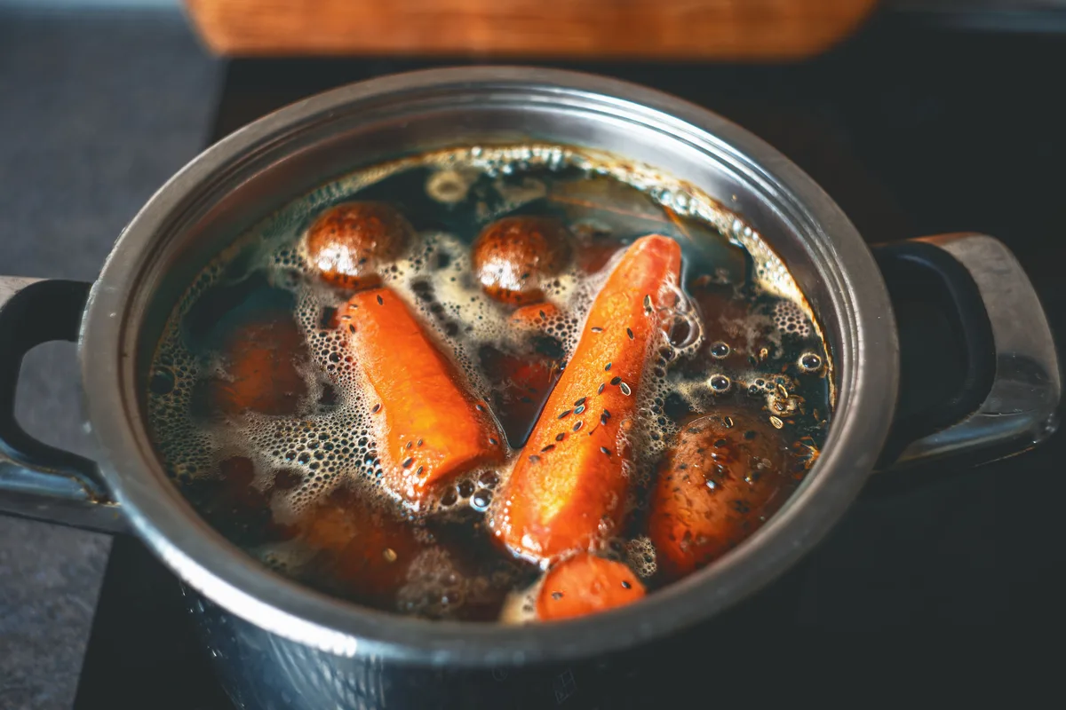 Whole carrots and other vegetables bubbling in a pot of dark stock with whole spices