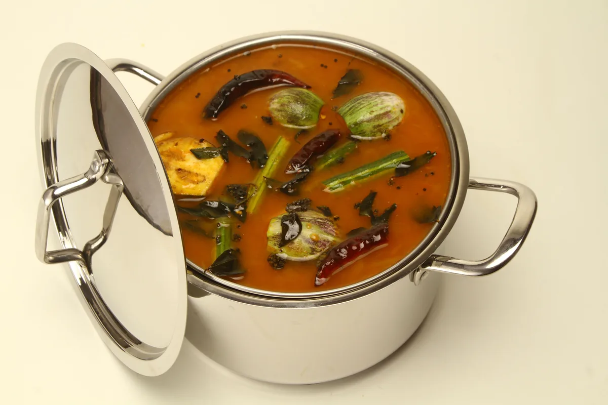 A steel pot of sambar with drumstick pods, small eggplants, curry leaves and dried red chilies