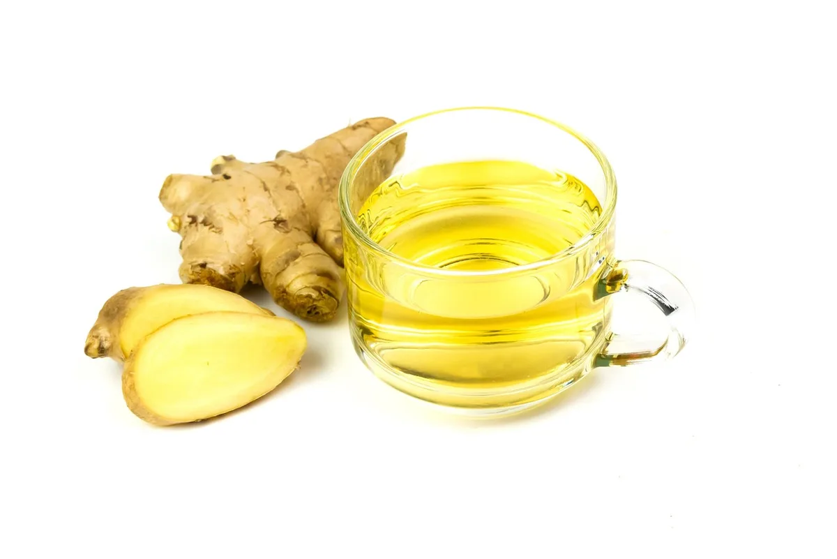 A glass cup of pale golden tea next to a fresh ginger root and two thick slices of ginger