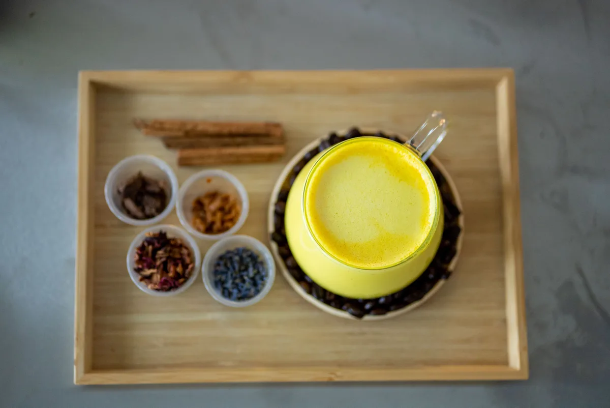 A glass of bright yellow golden milk on a wooden tray with cinnamon sticks and small cups of dried spices and flowers