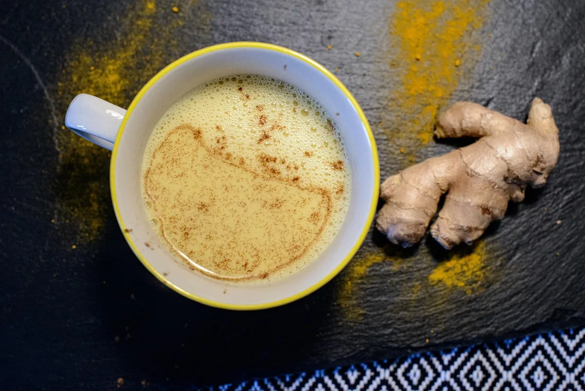A mug of frothy turmeric milk on dark slate, next to a knob of fresh ginger and a dusting of turmeric