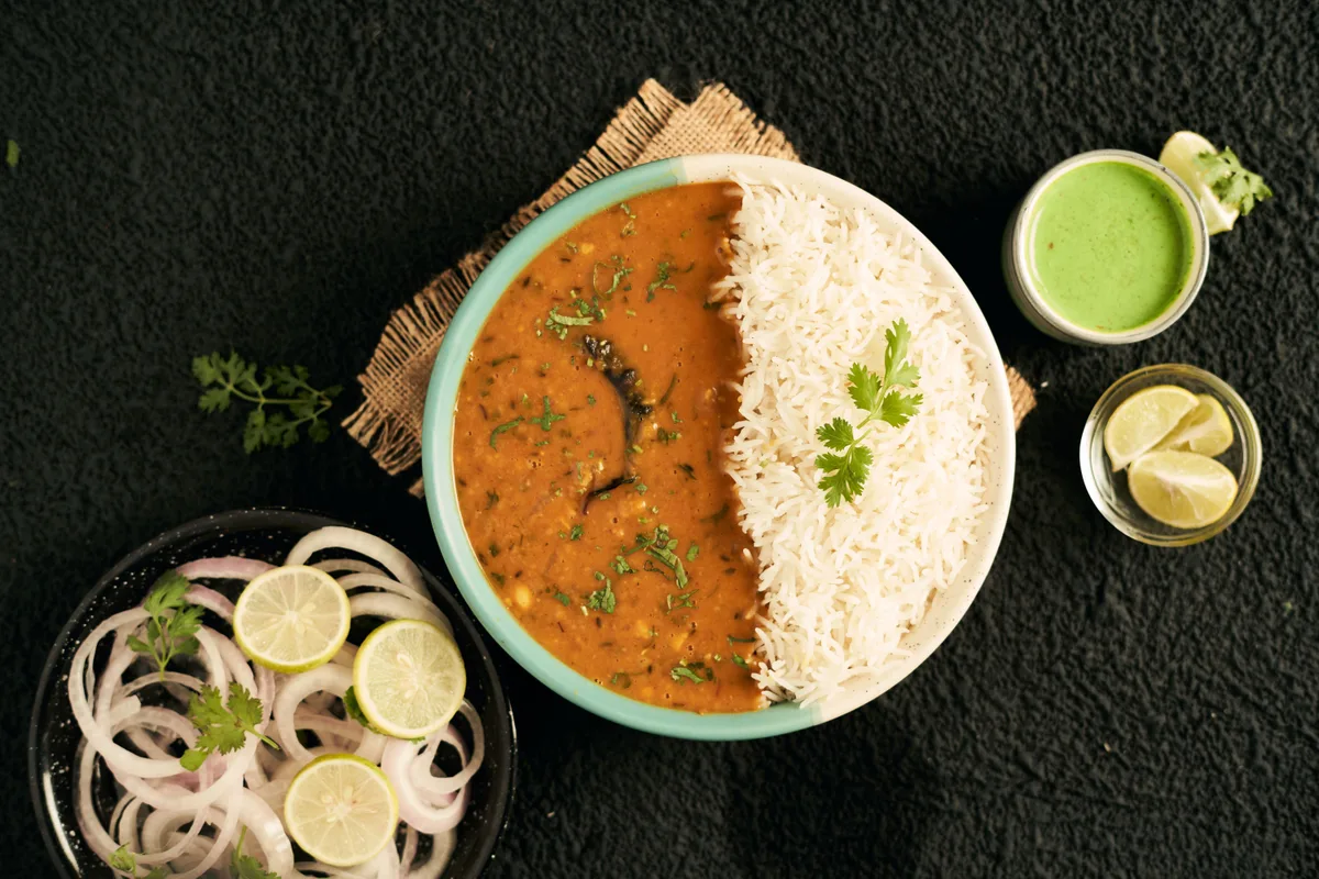 A bowl of dal and white rice with sliced onion, lime wedges and green chutney on the side