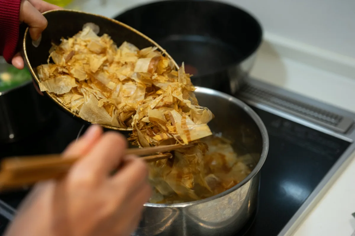 Dried bonito flakes being tipped from a bowl into a pot of hot water to make dashi