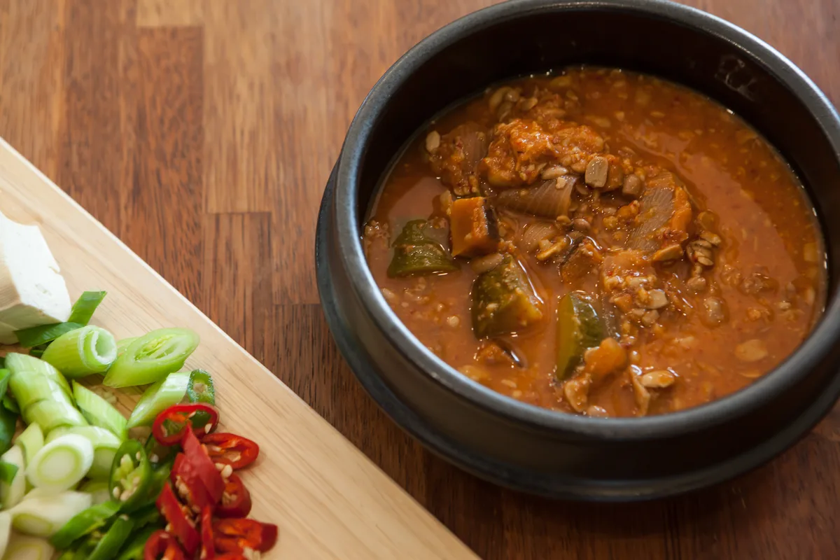 Doenjang jjigae in a black earthenware pot beside a board of sliced scallions, chilli and tofu