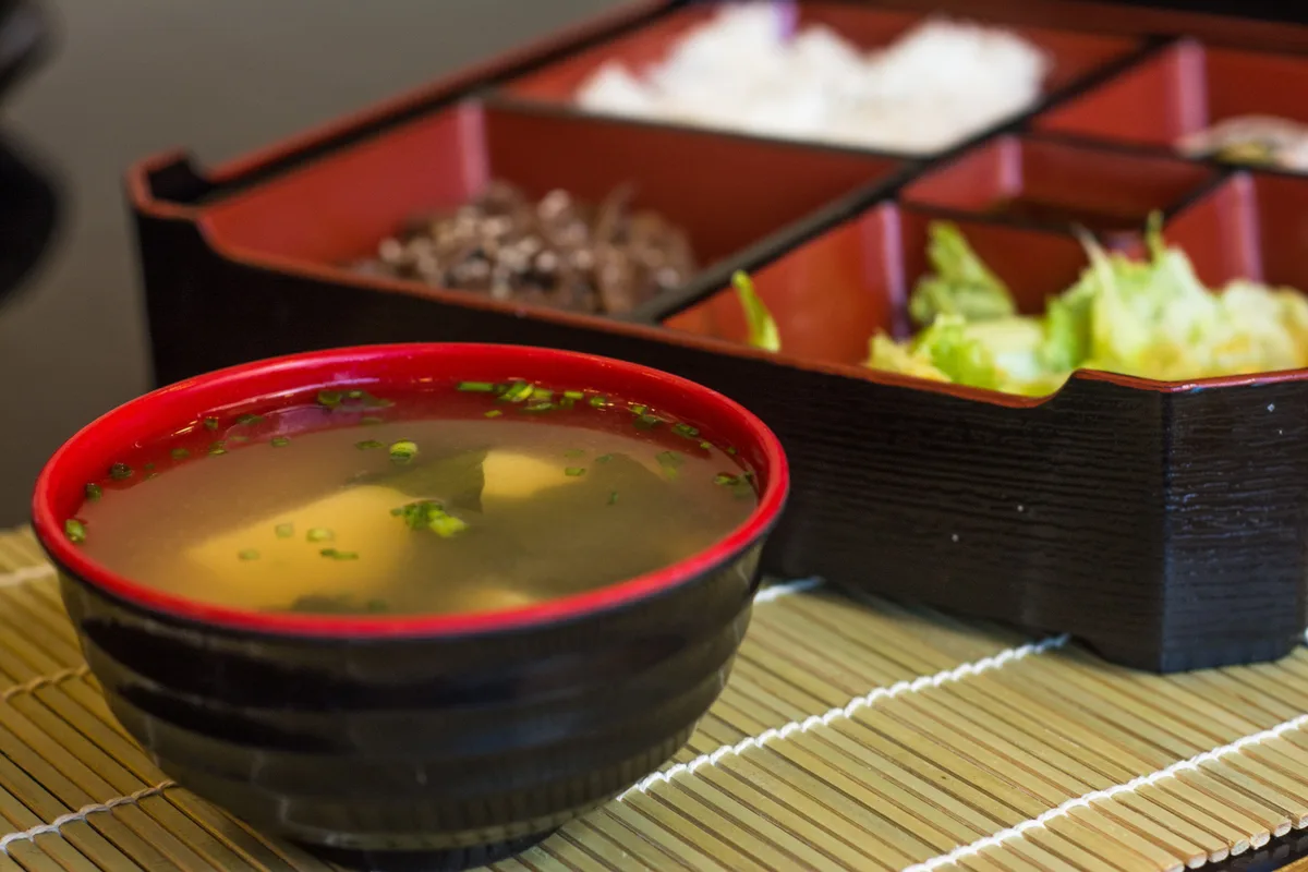 A bowl of pale miso soup with wakame in front of a lacquered bento box
