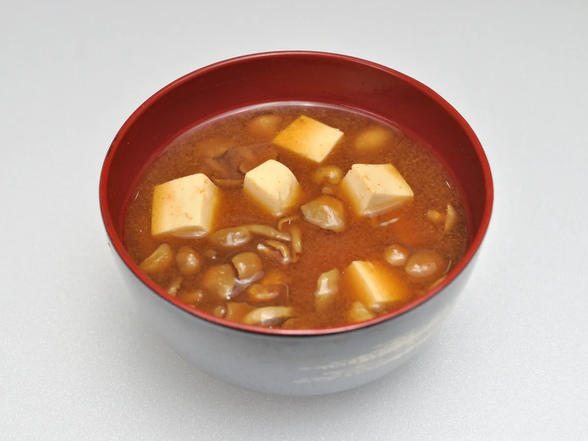 A dark reddish miso soup with tofu cubes and small nameko mushrooms in a lacquer bowl