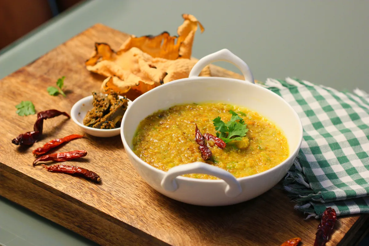 A bowl of yellow khichdi with dried red chillies and coriander, with papad and pickle on a wooden board