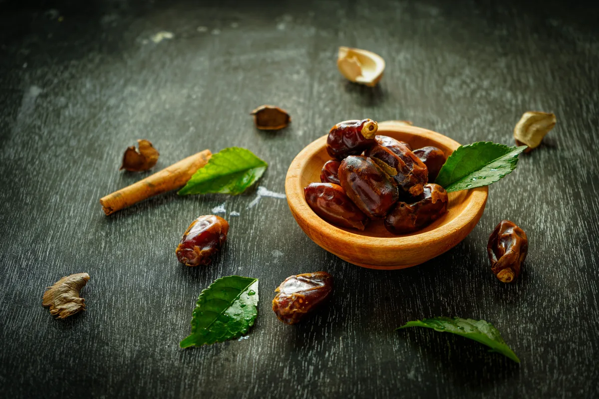 Dates in a small wooden bowl on a dark table, with a cinnamon stick and fresh leaves