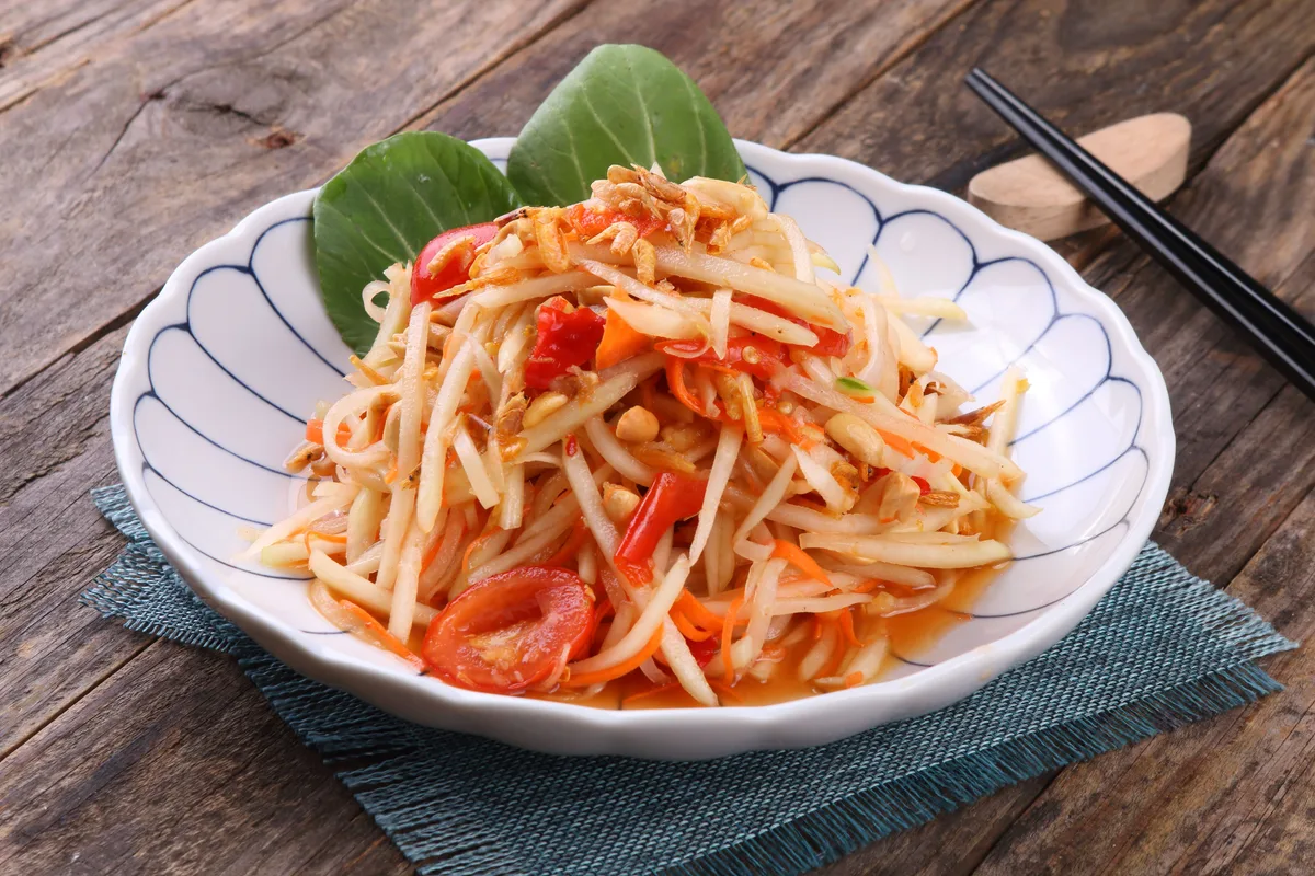Som tam, shredded green papaya salad with tomatoes, in a white bowl on a wooden table