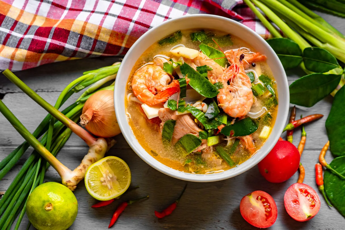 A bowl of Thai prawn soup surrounded by lemongrass stalks, lime leaves, lime, shallot and fresh chillies