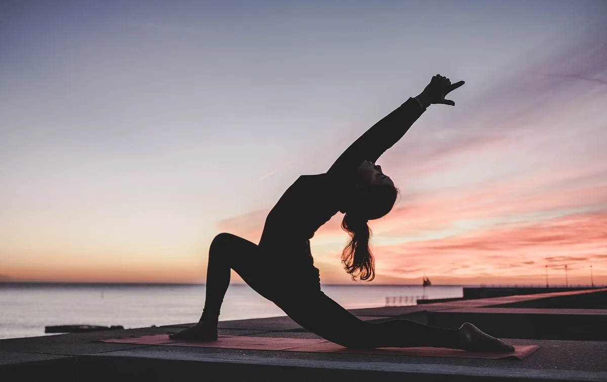The silhouette of a person in a deep lunge yoga pose against a pink sky by the sea