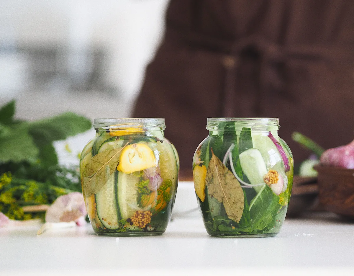 Two glass jars of vegetables in brine, with cucumber slices, bay leaves, mustard seeds and herbs