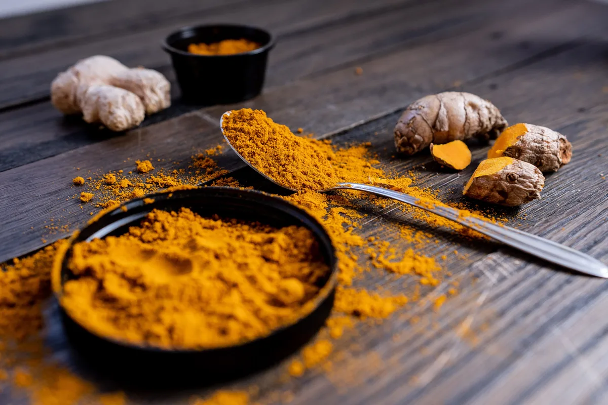 Ground turmeric in a black dish and on a spoon, with fresh turmeric and ginger roots on a dark wooden table