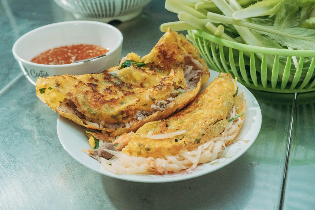 Crisp yellow bánh xèo crepes on a plate, with a bowl of dipping sauce and a basket of greens for wrapping