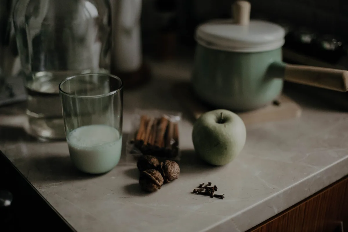 A glass of milk on a kitchen counter next to cinnamon sticks, walnuts, cloves and a green apple