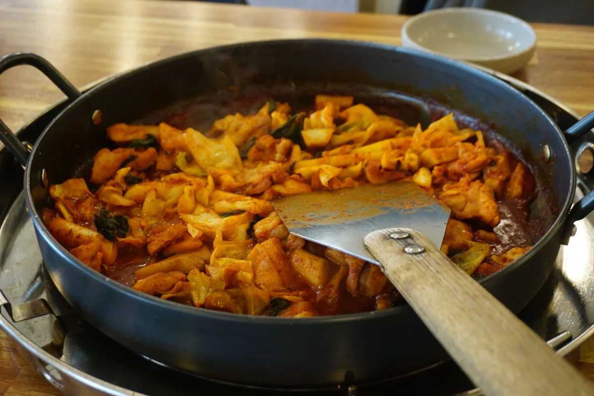 Dak galbi cooking down in red gochujang sauce in a wide pan, with a wooden spatula