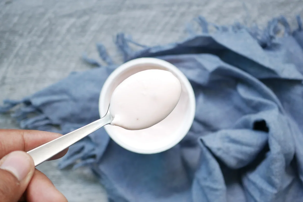 A hand lifting a spoonful of plain white yogurt from a small bowl on a blue cloth